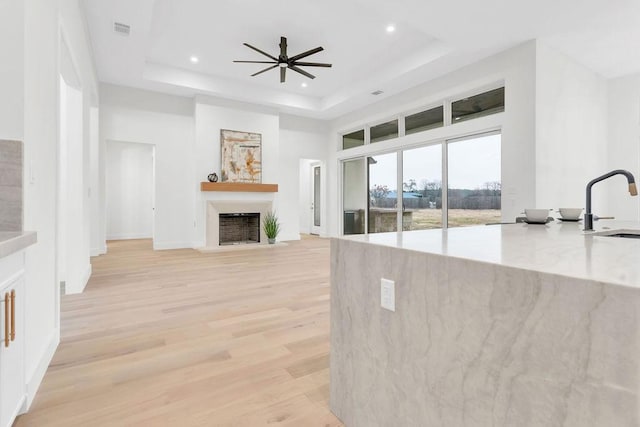 living room with ceiling fan, a towering ceiling, a tray ceiling, light stone countertops, and light wood-type flooring