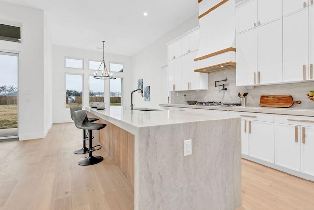 kitchen featuring a kitchen island with sink, backsplash, white cabinetry, hanging light fixtures, and light hardwood / wood-style floors