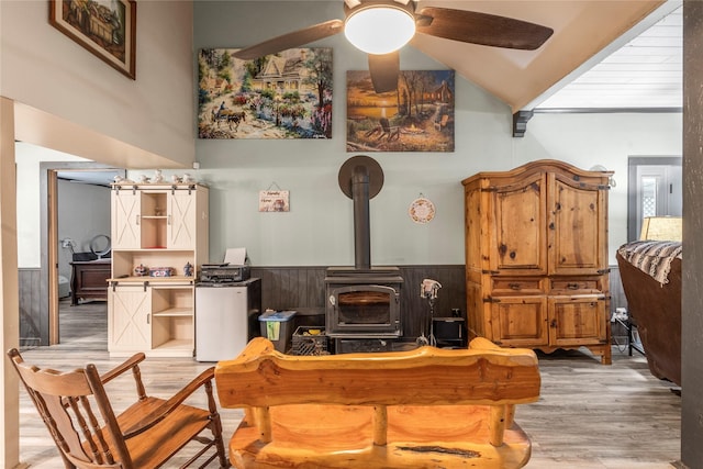 living room featuring wood finished floors, a wood stove, a ceiling fan, and wainscoting