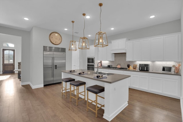 kitchen with sink, white cabinetry, built in appliances, an island with sink, and pendant lighting