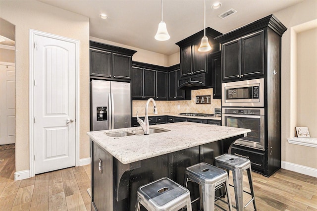 kitchen with sink, light stone counters, hanging light fixtures, appliances with stainless steel finishes, and an island with sink