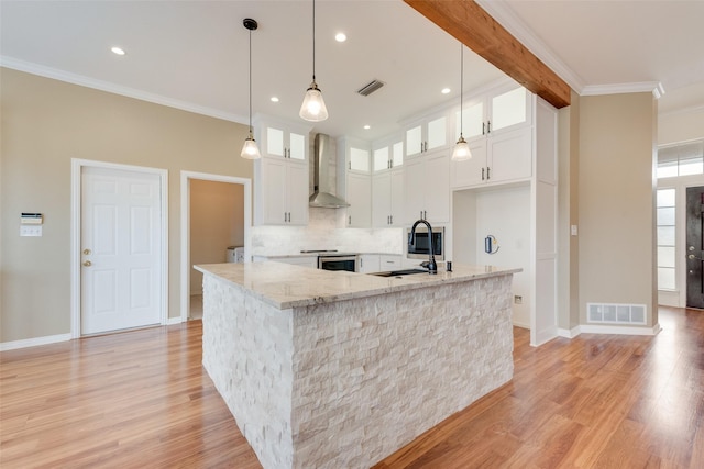 kitchen with wall chimney exhaust hood, white cabinetry, light stone counters, decorative light fixtures, and a center island with sink