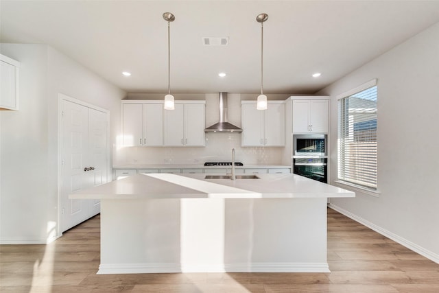 kitchen with white cabinets, decorative light fixtures, a center island with sink, and wall chimney range hood