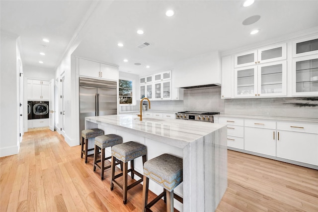 kitchen with a kitchen island with sink, light stone countertops, stainless steel built in fridge, white cabinets, and a kitchen bar