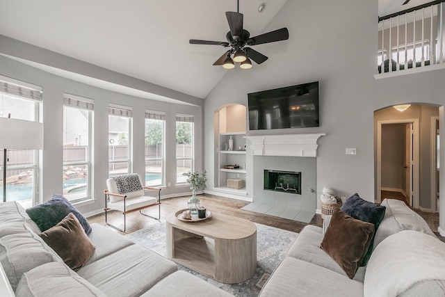 living room featuring high vaulted ceiling, light wood-type flooring, built in features, a tile fireplace, and ceiling fan