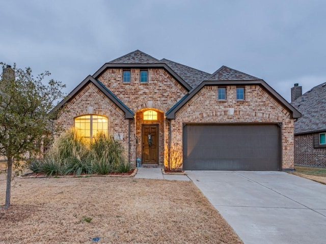 french provincial home featuring concrete driveway, brick siding, an attached garage, and roof with shingles