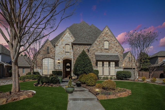 french country style house featuring stone siding, brick siding, roof with shingles, and a front yard