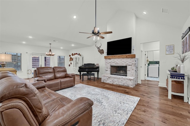 living room featuring ceiling fan with notable chandelier, high vaulted ceiling, washer / clothes dryer, hardwood / wood-style flooring, and a brick fireplace