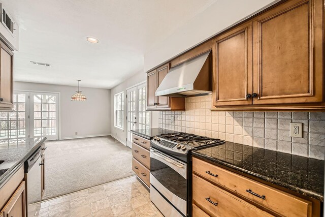 kitchen with wall chimney range hood, appliances with stainless steel finishes, tasteful backsplash, light colored carpet, and dark stone counters