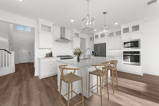 kitchen with pendant lighting, wall chimney range hood, a kitchen island with sink, white cabinetry, and stainless steel appliances