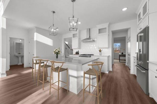 kitchen featuring a kitchen island with sink, high quality fridge, white cabinets, decorative light fixtures, and wall chimney exhaust hood