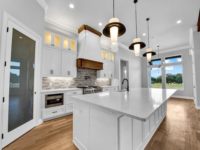 kitchen featuring sink, custom exhaust hood, white cabinetry, range, and a center island with sink