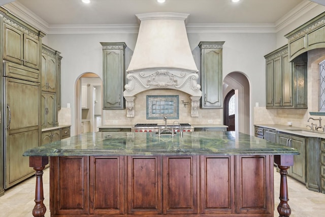 kitchen featuring paneled built in refrigerator, custom range hood, dark stone counters, and a center island with sink