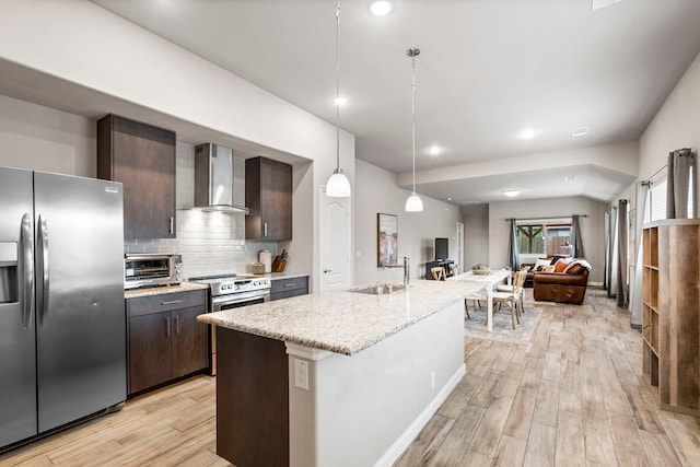 kitchen with wall chimney range hood, sink, a kitchen island with sink, hanging light fixtures, and stainless steel appliances