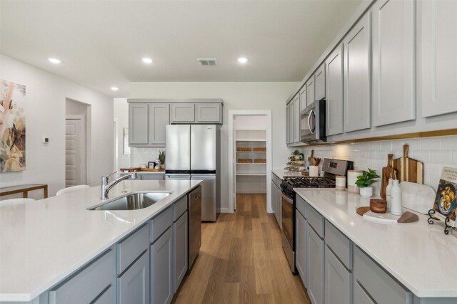 kitchen with sink, gray cabinetry, a center island with sink, light wood-type flooring, and appliances with stainless steel finishes