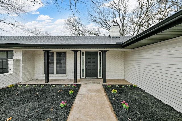 property entrance with a shingled roof, covered porch, brick siding, and a chimney