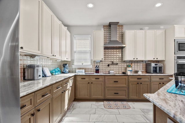 kitchen featuring light stone counters, wall chimney range hood, decorative backsplash, and appliances with stainless steel finishes