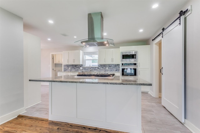 kitchen with stainless steel appliances, white cabinetry, island exhaust hood, dark stone counters, and a barn door