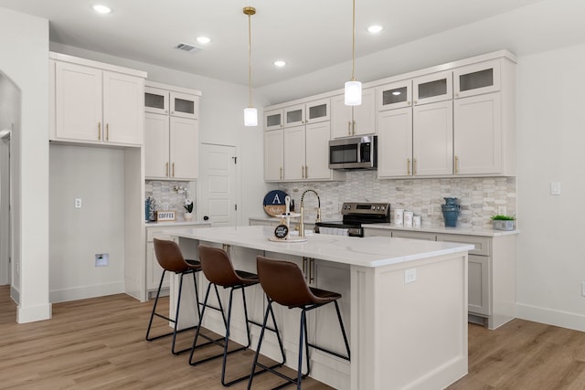 kitchen with white cabinetry, decorative light fixtures, appliances with stainless steel finishes, a kitchen island with sink, and light hardwood / wood-style floors