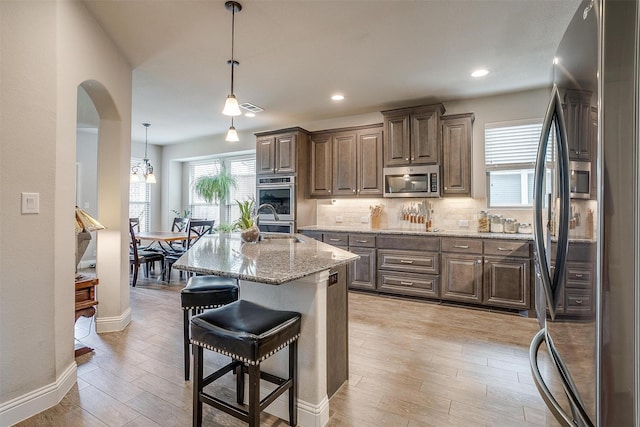 kitchen featuring decorative light fixtures, stainless steel appliances, light stone countertops, a kitchen island with sink, and decorative backsplash