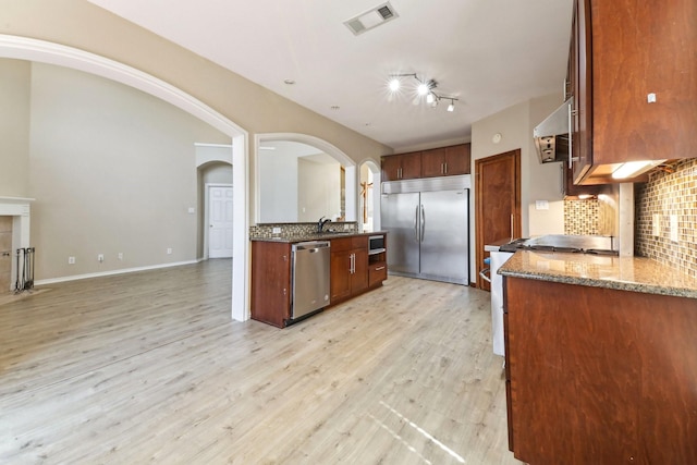 kitchen with dark stone countertops, light wood-type flooring, stainless steel appliances, range hood, and backsplash