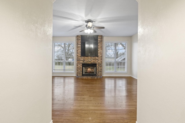 unfurnished living room featuring a brick fireplace, dark wood-type flooring, ceiling fan, and plenty of natural light