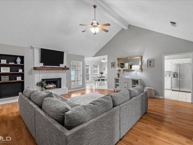 living room with a brick fireplace, ceiling fan, beamed ceiling, and light wood-type flooring