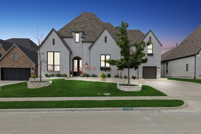french provincial home featuring concrete driveway, a shingled roof, a lawn, brick siding, and a garage
