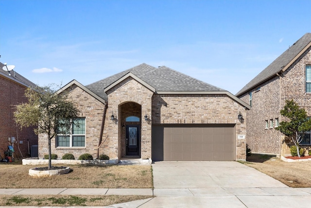 french country style house with roof with shingles, brick siding, an attached garage, central AC unit, and driveway