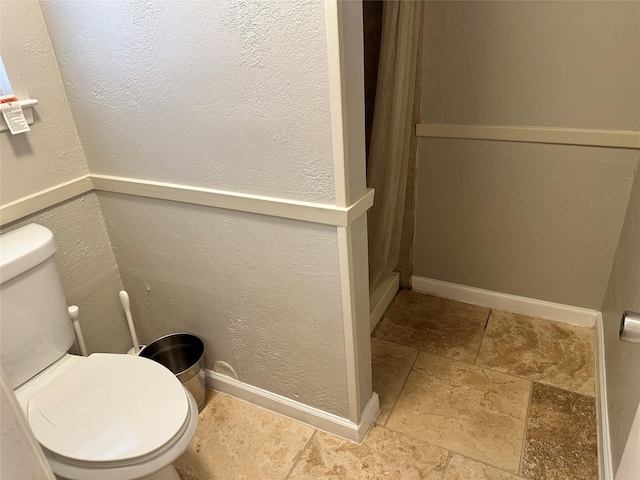 bathroom with toilet, stone finish flooring, baseboards, and a textured wall