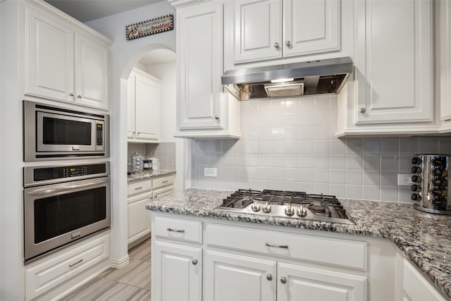 kitchen featuring appliances with stainless steel finishes, range hood, white cabinetry, decorative backsplash, and light stone counters