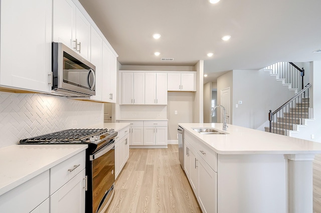 kitchen with sink, appliances with stainless steel finishes, a kitchen island with sink, white cabinetry, and light wood-type flooring