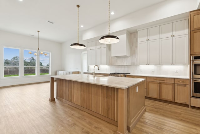 kitchen featuring sink, white cabinetry, hanging light fixtures, a kitchen island with sink, and wall chimney range hood