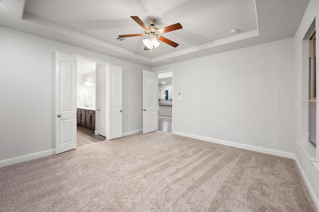 unfurnished bedroom with ceiling fan, ensuite bath, a tray ceiling, and light carpet