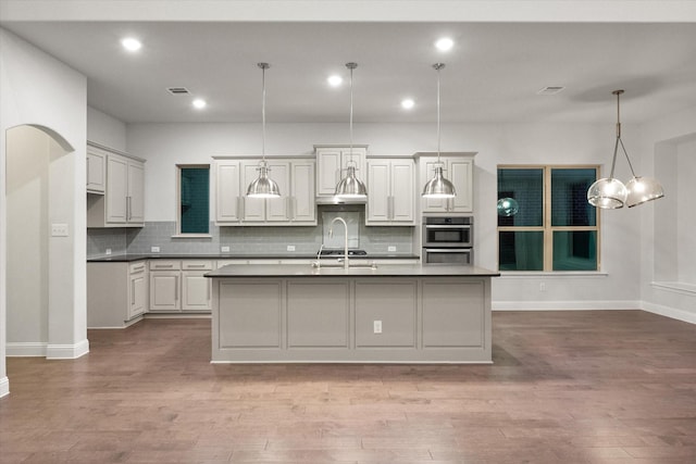 kitchen with tasteful backsplash, wood-type flooring, hanging light fixtures, a center island with sink, and double oven