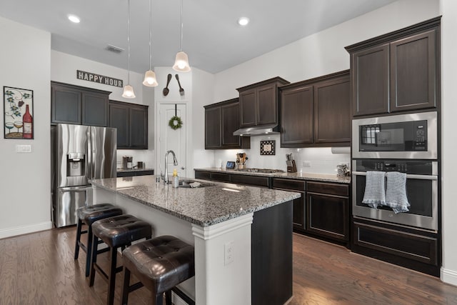 kitchen featuring appliances with stainless steel finishes, decorative light fixtures, sink, dark stone counters, and a kitchen island with sink