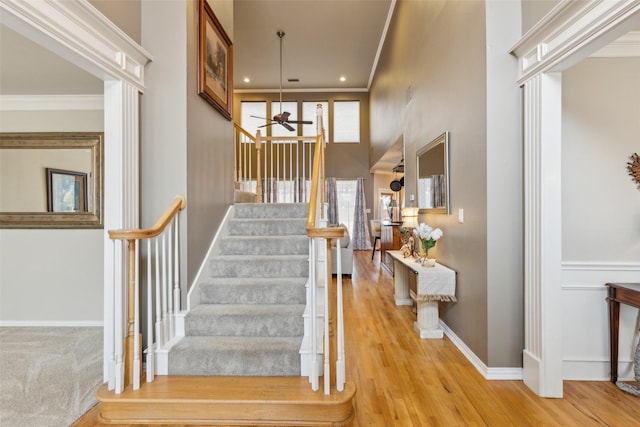 stairway with a towering ceiling, ornamental molding, a ceiling fan, wood finished floors, and baseboards