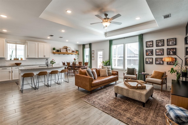 living room with sink, crown molding, light hardwood / wood-style flooring, a raised ceiling, and ceiling fan