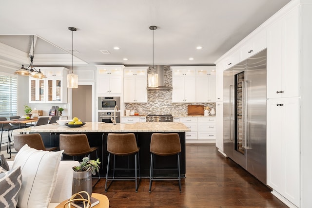 kitchen featuring built in refrigerator, light stone countertops, white cabinets, decorative light fixtures, and wall chimney exhaust hood