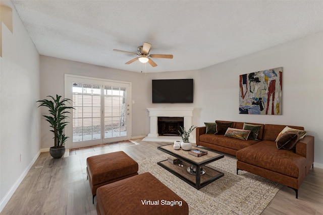 living area featuring a fireplace with raised hearth, ceiling fan, a textured ceiling, baseboards, and light wood-type flooring