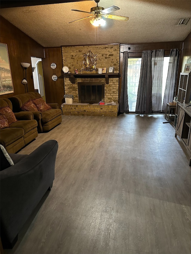 living room featuring ceiling fan, wood-type flooring, a textured ceiling, a brick fireplace, and wood walls