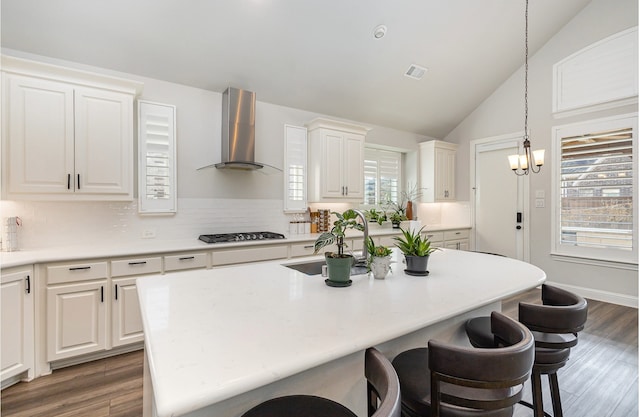 kitchen with decorative light fixtures, white cabinets, stainless steel gas cooktop, a kitchen island with sink, and wall chimney exhaust hood