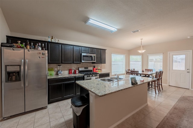 kitchen featuring sink, a kitchen island with sink, stainless steel appliances, light stone counters, and decorative light fixtures