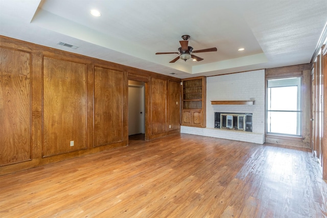 unfurnished living room featuring a ceiling fan, visible vents, a brick fireplace, light wood-style floors, and a tray ceiling