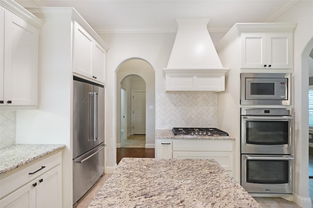 kitchen featuring appliances with stainless steel finishes, ornamental molding, white cabinets, light stone countertops, and premium range hood