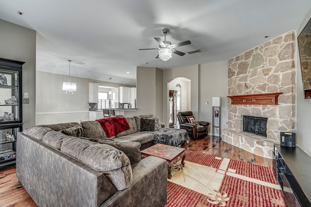 living room featuring visible vents, ceiling fan with notable chandelier, wood finished floors, arched walkways, and a stone fireplace