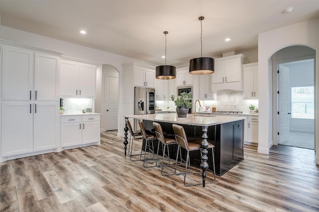 kitchen with pendant lighting, a kitchen island with sink, stainless steel appliances, white cabinets, and light wood-type flooring