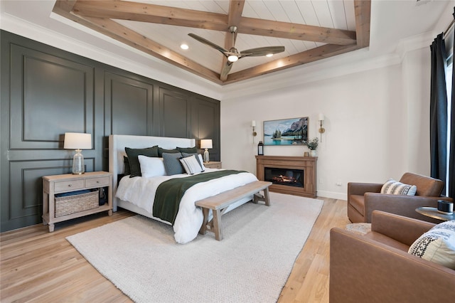 bedroom featuring a glass covered fireplace, ceiling fan, light wood-style flooring, beamed ceiling, and a decorative wall