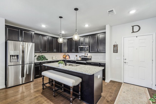 kitchen with appliances with stainless steel finishes, a kitchen island with sink, hanging light fixtures, a kitchen breakfast bar, and light stone countertops