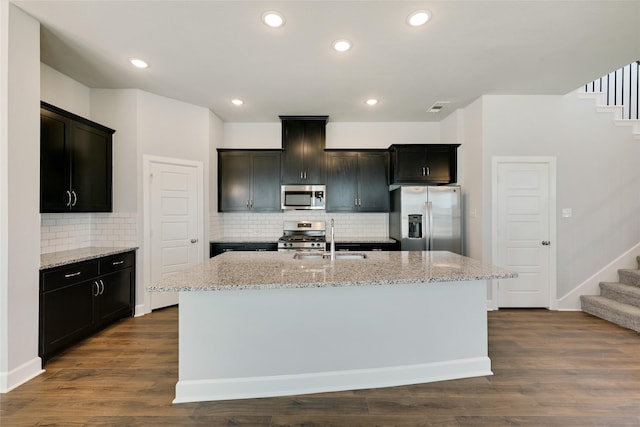 kitchen with appliances with stainless steel finishes, sink, light stone countertops, and an island with sink